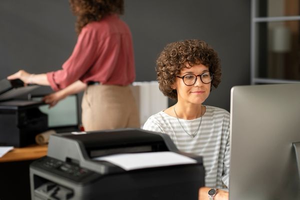 A women is sitting at her workstation beside a desktop printer. A colleague is standing at their workstation in the background using a printer.